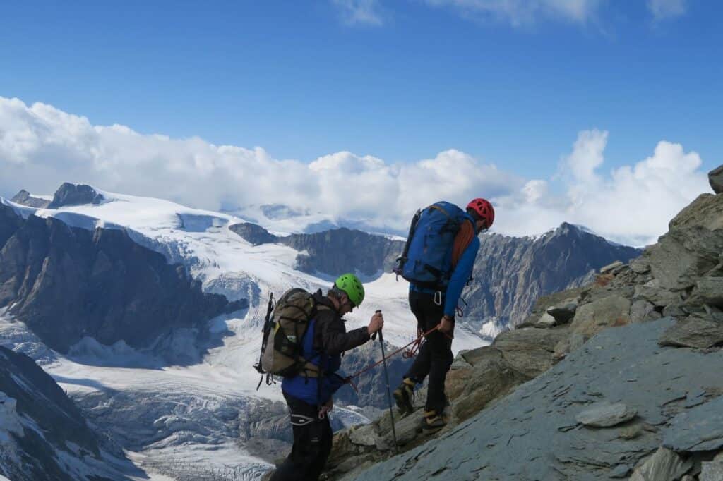 Mountain guide and mountaineering ascending the top of a mountain looking over to large glacier in the background and peaks of other mountains.